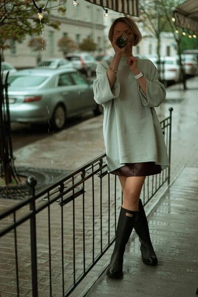 A young woman in the rain on an open terrace drinks tea from a large glass