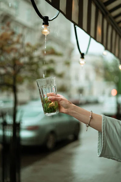 オープンテラスで雨の中の若い女性が大きなガラスからお茶を飲む — ストック写真