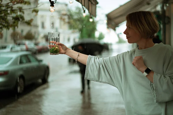 オープンテラスで雨の中の若い女性が大きなガラスからお茶を飲む — ストック写真