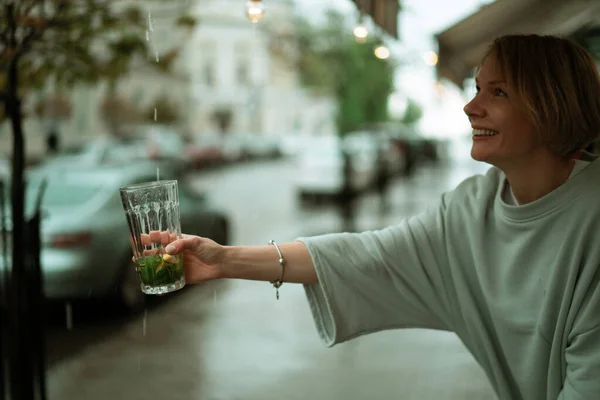 オープンテラスで雨の中の若い女性が大きなガラスからお茶を飲む — ストック写真