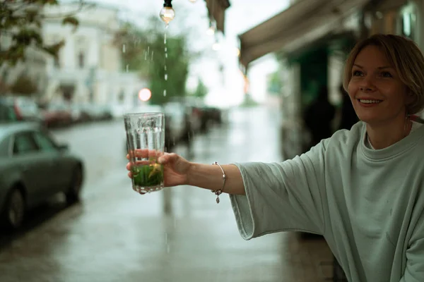 オープンテラスで雨の中の若い女性が大きなガラスからお茶を飲む — ストック写真