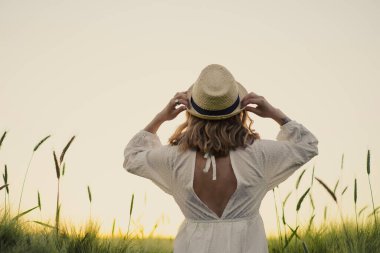 Sarı uzun saçlı, beyaz elbiseli, buğday tarlasında hasır şapkalı güzel bir kadın. Güneşte uçan saçlar, yaz. Hayalperestlerin zamanı, altın gün batımı.