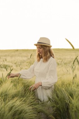 Sarı uzun saçlı, beyaz elbiseli, buğday tarlasında hasır şapkalı güzel bir kadın. Güneşte uçan saçlar, yaz. Hayalperestlerin zamanı, altın gün batımı.