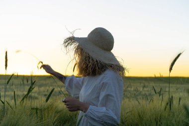Sarı uzun saçlı, beyaz elbiseli, buğday tarlasında hasır şapkalı güzel bir kadın. Güneşte uçan saçlar, yaz. Hayalperestlerin zamanı, altın gün batımı.