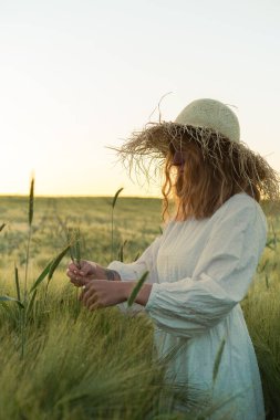 Sarı uzun saçlı, hasır şapkalı beyaz elbiseli güzel bir kadın buketi buğday tarlasında topluyor. Güneşte uçan saçlar, yaz. Hayalperestlerin zamanı, altın gün batımı.