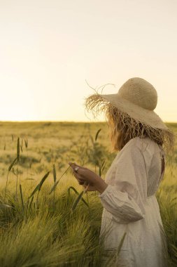 Sarı uzun saçlı, beyaz elbiseli, buğday tarlasında hasır şapkalı güzel bir kadın. Güneşte uçan saçlar, yaz. Hayalperestlerin zamanı, altın gün batımı.