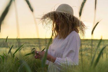 Sarı uzun saçlı, hasır şapkalı beyaz elbiseli güzel bir kadın buketi buğday tarlasında topluyor. Güneşte uçan saçlar, yaz. Hayalperestlerin zamanı, altın gün batımı.