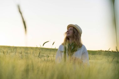 Sarı uzun saçlı, hasır şapkalı beyaz elbiseli güzel bir kadın buketi buğday tarlasında topluyor. Güneşte uçan saçlar, yaz. Hayalperestlerin zamanı, altın gün batımı.