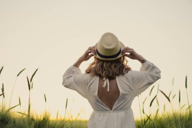 Sarı uzun saçlı, beyaz elbiseli, buğday tarlasında hasır şapkalı güzel bir kadın. Güneşte uçan saçlar, yaz. Hayalperestlerin zamanı, altın gün batımı.