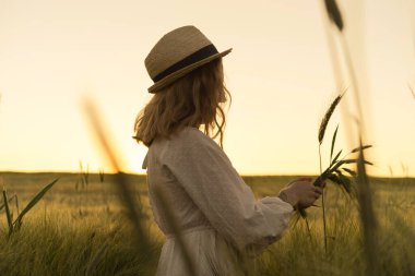 Sarı uzun saçlı, hasır şapkalı beyaz elbiseli güzel bir kadın buketi buğday tarlasında topluyor. Güneşte uçan saçlar, yaz. Hayalperestlerin zamanı, altın gün batımı.