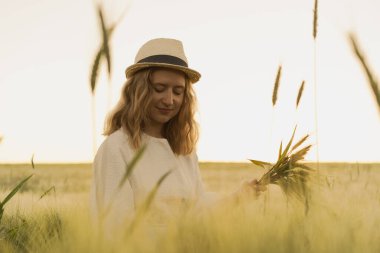 Sarı uzun saçlı, hasır şapkalı beyaz elbiseli güzel bir kadın buketi buğday tarlasında topluyor. Güneşte uçan saçlar, yaz. Hayalperestlerin zamanı, altın gün batımı.