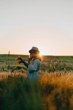 Sarı uzun saçlı, beyaz elbiseli, buğday tarlasında hasır şapkalı güzel bir kadın. Güneşte uçan saçlar, yaz. Hayalperestlerin zamanı, altın gün batımı.
