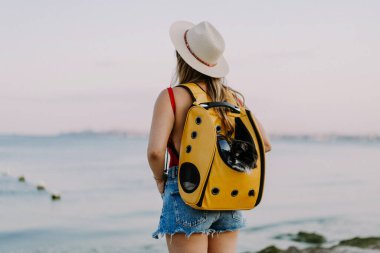 Deniz kıyısında sırt çantasında kedisi olan genç bir kadın. Bir evcil hayvanla seyahat konsepti.