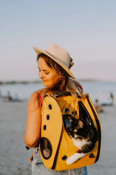 young woman with a cat in a backpack on the seashore. Travel concept with a pet.