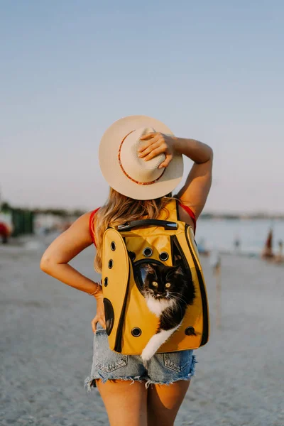 young woman with a cat in a backpack on the seashore. Travel concept with a pet.