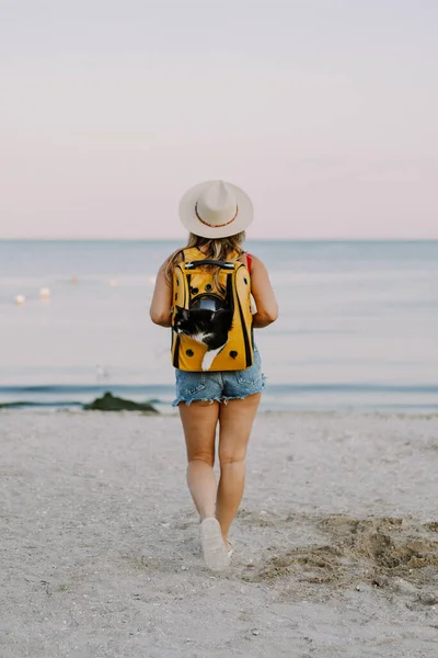 young woman with a cat in a backpack on the seashore. Travel concept with a pet.