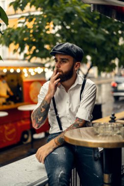 Caddedeki bir kafede sigara içen sakallı genç bir adam. Şehirde beyaz gömlekli ve pantolon askısı takan romantik bir adam. Peaky Blinders. eski moda retro.