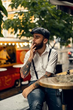 Caddedeki bir kafede sigara içen sakallı genç bir adam. Şehirde beyaz gömlekli ve pantolon askısı takan romantik bir adam. Peaky Blinders. eski moda retro.