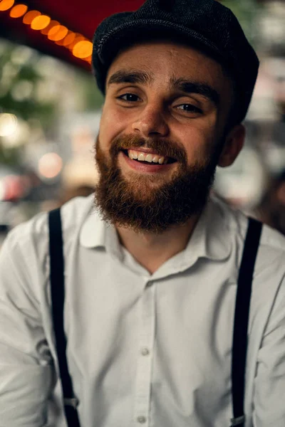 Young bearded man in a cafe on the street with a glass of wine. Romantic guy in a white shirt cap and suspenders in the city. Peaky Blinders. old fassion retro.