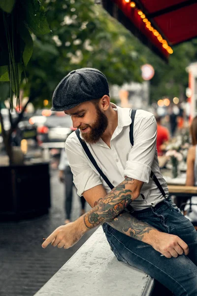 Young bearded man in a cafe on the street smokes a cigarette. Romantic guy in a white shirt cap and suspenders in the city. Peaky Blinders. old fashionable retro.