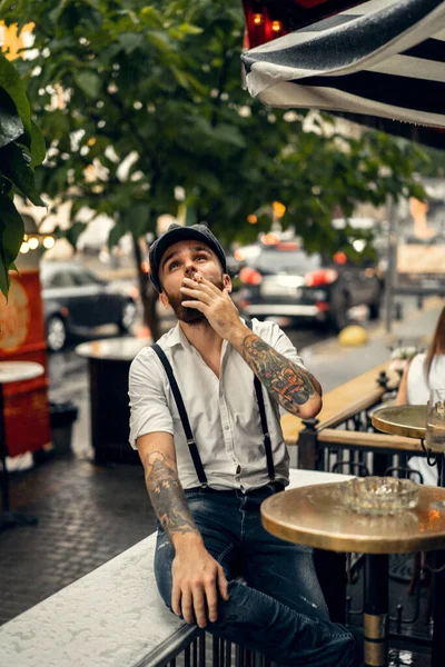 Young bearded man in a cafe on the street smokes a cigarette. Romantic guy in a white shirt cap and suspenders in the city. Peaky Blinders. old fashionable retro.