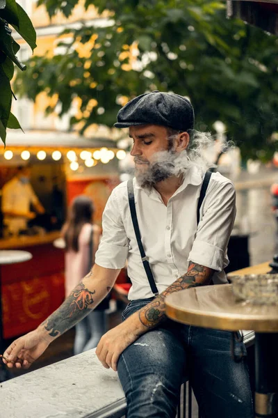 Young bearded man in a cafe on the street smokes a cigarette. Romantic guy in a white shirt cap and suspenders in the city. Peaky Blinders. old fashionable retro.