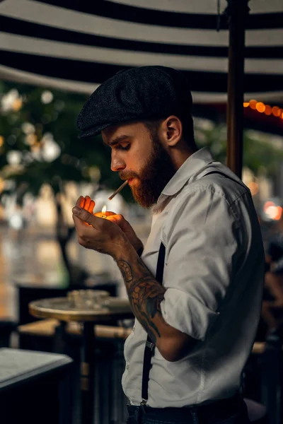 Young bearded man in a cafe on the street smokes a cigarette. Romantic guy in a white shirt cap and suspenders in the city. Peaky Blinders. old fashionable retro.