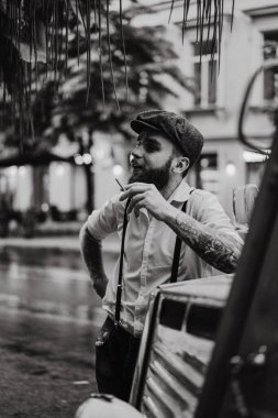 Caddedeki bir kafede sigara içen sakallı genç bir dövmeli adam. Şehirde beyaz gömlekli ve pantolon askısı takan romantik bir adam. Peaky Blinders. eski moda retro.