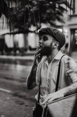 Caddedeki bir kafede sigara içen sakallı genç bir dövmeli adam. Şehirde beyaz gömlekli ve pantolon askısı takan romantik bir adam. Peaky Blinders. eski moda retro.