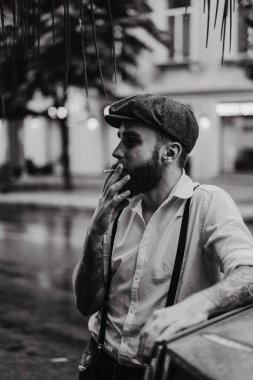 Caddedeki bir kafede sigara içen sakallı genç bir dövmeli adam. Şehirde beyaz gömlekli ve pantolon askısı takan romantik bir adam. Peaky Blinders. eski moda retro.