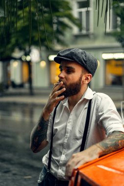 Caddedeki bir kafede sigara içen sakallı genç bir dövmeli adam. Şehirde beyaz gömlekli ve pantolon askısı takan romantik bir adam. Peaky Blinders. eski moda retro.