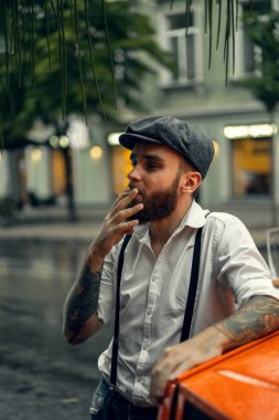 Caddedeki bir kafede sigara içen sakallı genç bir dövmeli adam. Şehirde beyaz gömlekli ve pantolon askısı takan romantik bir adam. Peaky Blinders. eski moda retro.