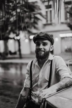 Caddedeki bir kafede sigara içen sakallı genç bir dövmeli adam. Şehirde beyaz gömlekli ve pantolon askısı takan romantik bir adam. Peaky Blinders. eski moda retro.