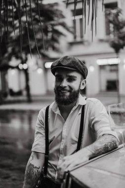 Caddedeki bir kafede sigara içen sakallı genç bir dövmeli adam. Şehirde beyaz gömlekli ve pantolon askısı takan romantik bir adam. Peaky Blinders. eski moda retro.