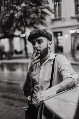 Caddedeki bir kafede sigara içen sakallı genç bir dövmeli adam. Şehirde beyaz gömlekli ve pantolon askısı takan romantik bir adam. Peaky Blinders. eski moda retro.