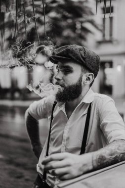 Caddedeki bir kafede sigara içen sakallı genç bir dövmeli adam. Şehirde beyaz gömlekli ve pantolon askısı takan romantik bir adam. Peaky Blinders. eski moda retro.