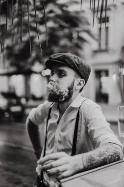 Caddedeki bir kafede sigara içen sakallı genç bir dövmeli adam. Şehirde beyaz gömlekli ve pantolon askısı takan romantik bir adam. Peaky Blinders. eski moda retro.