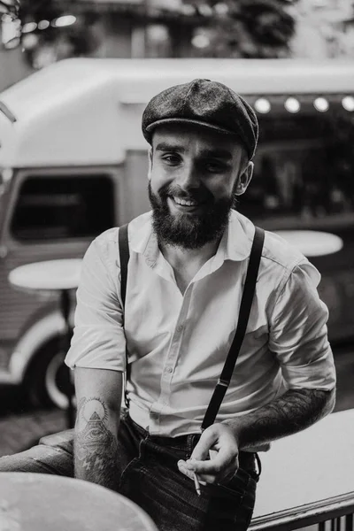 Young bearded man in a cafe on the street smokes a cigarette. Romantic guy in a white shirt cap and suspenders in the city. Peaky Blinders. old fashionable retro.