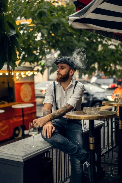 Young bearded man in a cafe on the street with a glass of wine. Romantic guy in a white shirt cap and suspenders in the city. Peaky Blinders. old fassion retro.