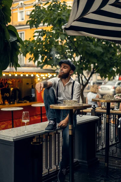 Young bearded man in a cafe on the street with a glass of wine. Romantic guy in a white shirt cap and suspenders in the city. Peaky Blinders. old fassion retro.