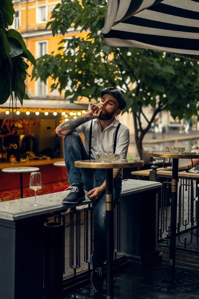 Young bearded man in a cafe on the street with a glass of wine. Romantic guy in a white shirt cap and suspenders in the city. Peaky Blinders. old fassion retro.