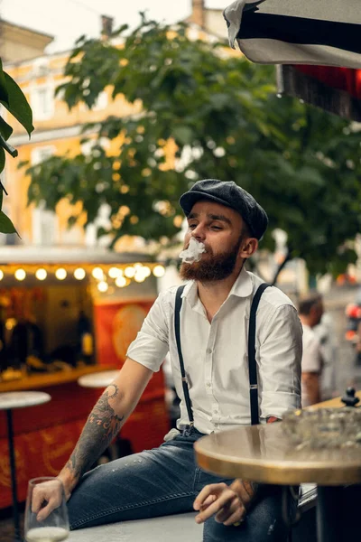 Young bearded man in a cafe on the street with a glass of wine. Romantic guy in a white shirt cap and suspenders in the city. Peaky Blinders. old fassion retro.