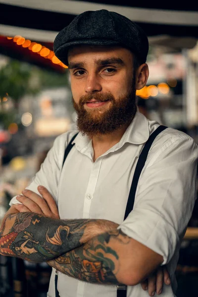 Young bearded man in a cafe on the street with a glass of wine. Romantic guy in a white shirt cap and suspenders in the city. Peaky Blinders. old fassion retro.