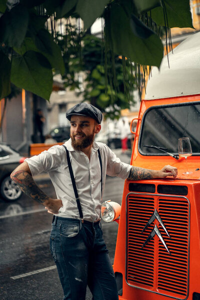 Young bearded man in a cafe on the street with a glass of wine. Romantic guy in a white shirt cap and suspenders in the city. Peaky Blinders. old fassion retro.