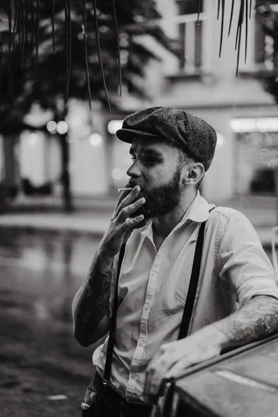 Young bearded tattoord man in a cafe on the street smokes a cigarette. Romantic guy in a white shirt cap and suspenders in the city. Peaky Blinders. old fashionable retro.