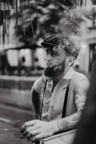 街上一家咖啡馆里的一个留着胡子的纹身男子吸烟 城市里穿着白衬衫帽和吊袜带的浪漫男人 小眼镜片 旧式复古风格 — 图库照片