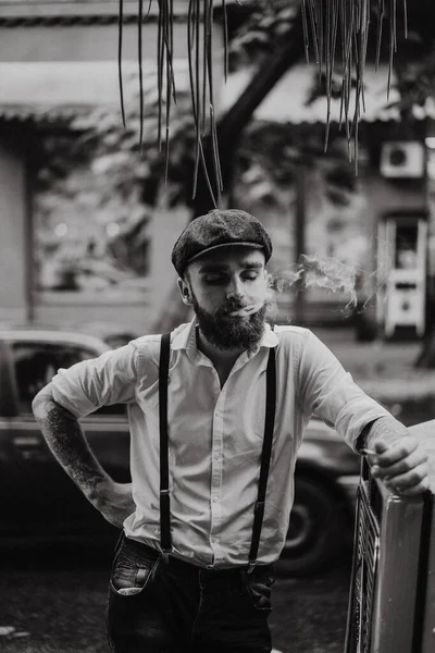 Young bearded tattoord man in a cafe on the street smokes a cigarette. Romantic guy in a white shirt cap and suspenders in the city. Peaky Blinders. old fashionable retro.