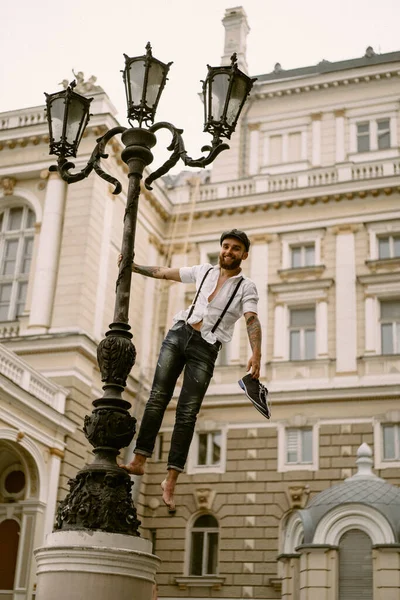 Young bearded tattooed man. A romantic guy in a white shirt, cap and suspenders walks in the city. revolves around a lamppost. Peaky Blinders. old-fashioned, retro.