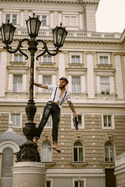 Young bearded tattooed man. A romantic guy in a white shirt, cap and suspenders walks in the city. revolves around a lamppost. Peaky Blinders. old-fashioned, retro.