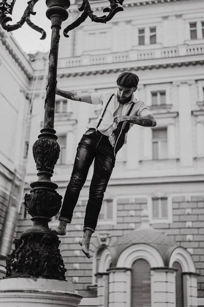 Young bearded tattooed man. A romantic guy in a white shirt, cap and suspenders walks in the city. revolves around a lamppost. Peaky Blinders. old-fashioned, retro.